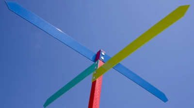 Vier farbige Metallstreben in Blau, Grün, Rot und Gelb, die an einem roten Pfosten befestigt sind, vor blauem Himmel.