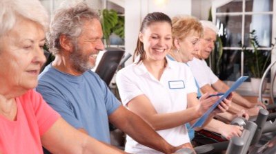 Mehrere Personen sitzen nebeneinander auf Fitnessgeräten, eine trägt weiße Arbeitskleidung und hält ein Klemmbrett.