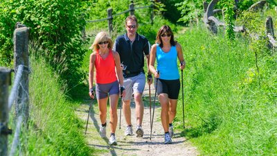 Drei Personen Wandern im Freien auf einem schmalen Weg der umgeben von hohem Gras ist.