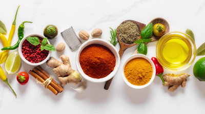 Verschiedene Gewürze und Zutaten, darunter Zimtstangen, Ingwer, Limetten, Chili, Kurkuma, Paprikapulver, Olivenöl und frische Kräuter, auf weißem Untergrund angeordnet.