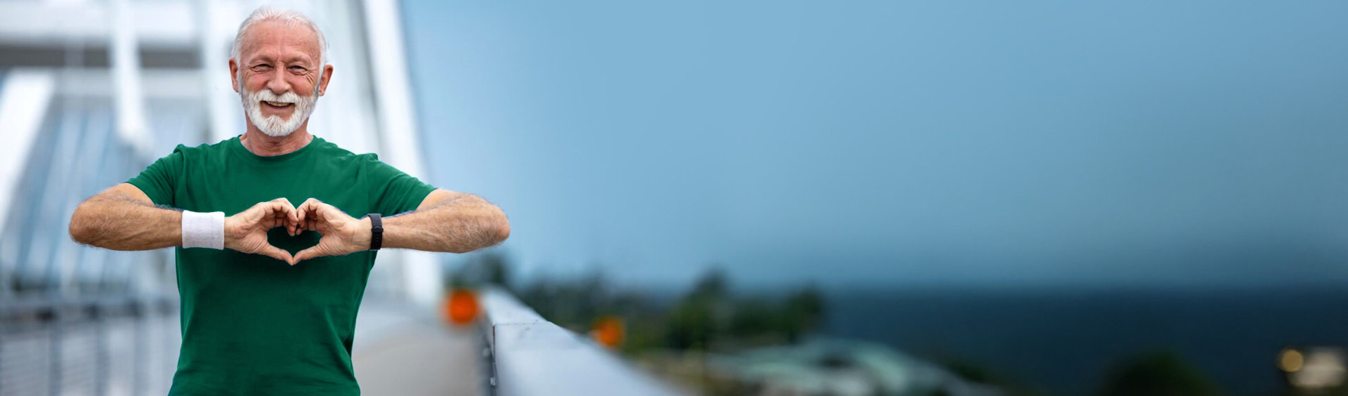 Ein Mann in grünem T-Shirt steht auf einer Brücke und formt mit seinen Händen ein Herz. Im Hintergrund ist unscharf das Meer und ein bewölkter Himmel zu sehen.