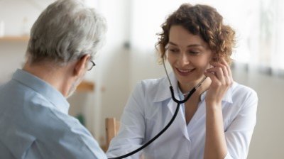 Ärztin mit weißem Kittel hört mit Stethoskop den Rücken eines Mannes ab.