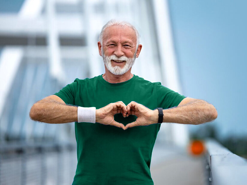 Ein Mann in grünem T-Shirt steht auf einer Brücke und formt mit seinen Händen ein Herz. Im Hintergrund ist unscharf das Meer und ein bewölkter Himmel zu sehen.