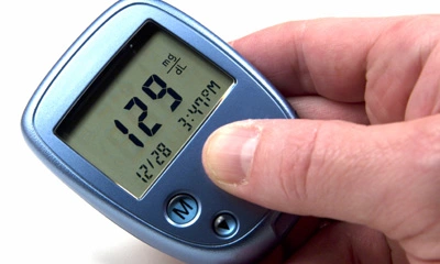 Hand hält blauen Blutzuckermessgerät mit Anzeige „129 mg/dL“, Datum „12/28“ und Uhrzeit „3:49 PM“.